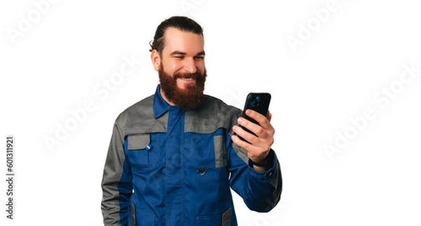 Fototapeta Handsome smiling young handyman in uniform has got a new client over the phone he is holding.