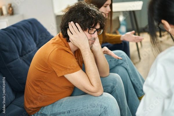 Fototapeta Couple having therapy session with a psychologist to solve their relationship problems and crisis. Sad man covering his ears with hands to avoid hearing abuse