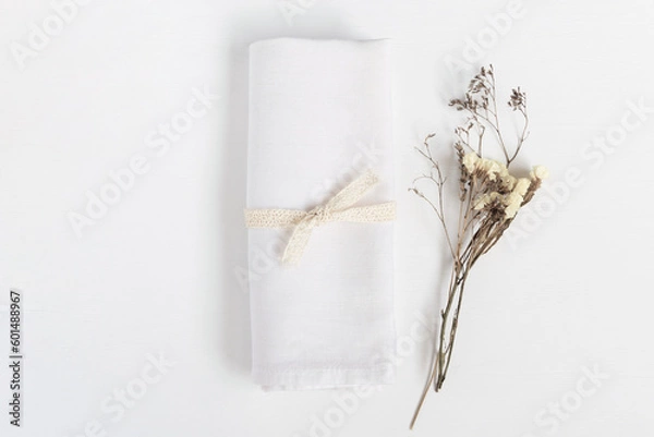 Fototapeta Linen napkin tied with ribbon on white table. Fabric and dried flowers in rustic style for mock up.