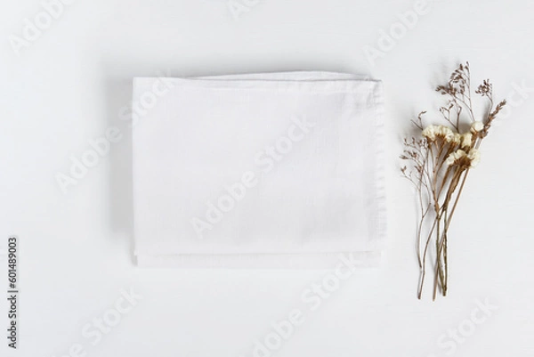 Fototapeta Stack of fabrics with dry flowers over light background. Flat lay with white folded linen cloth for mock up.