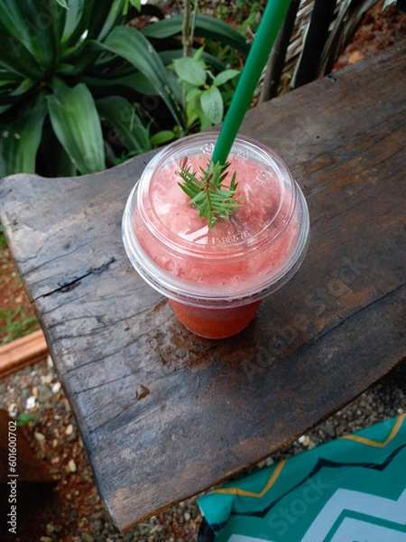 Fototapeta Strawberry smoothie in a plastic cup on a wooden table and green leaf in the garden of a roadside cafe. Top view. 
