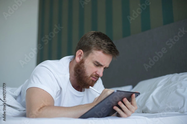 Fototapeta Sad young man drawing tablet screen, graphic designer work use write draw stylus pc pen on tablet computer lying on bed, sofa at home indoor flat, hobby weekends