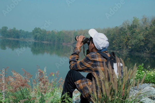 Obraz Asian boy in plaid shirt wears white cap, holding binoculars, standing on reservoir ridge during summer vacation and birdwatching activity, soft and selective focus, nature study and hobby concept.