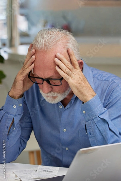 Fototapeta Shocked and frustrated senior man sitting at the table with laptop and looking at the bills and receipts. Elderly man struggling with finances calculating.