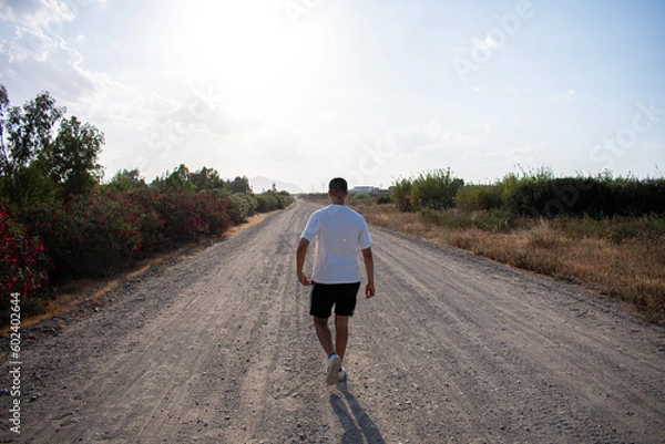 Fototapeta Boy walking on a dirt path in a field. Man walking along a path. Moroccan boy taking a stroll. Man walking in a field wearing a white t-shirt. Boy walking calmly through the countryside.