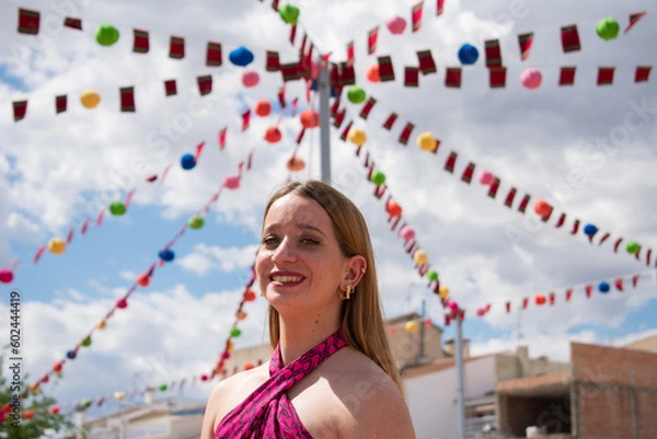 Obraz Blonde girl smiling. Happy young Caucasian woman dressed in a pink top, smiling joyfully, showing her happiness and well-being to someone. Body language. Young girl smiling at a festival. Party.
