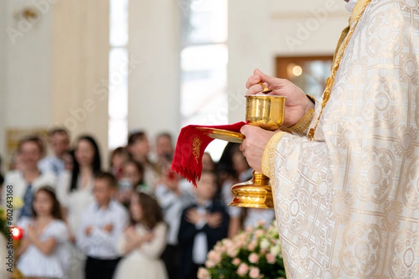Fototapeta A priest of the Catholic Church stands in front of the parishioners during the festive Holy Liturgy, holding the chalice with the sacraments in his hands. First Confession and solemn Holy Communion