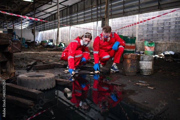 Fototapeta Worker or scientist industry wearing protective safety uniform, white glove and gas mask under cleaning up and lapping spilled oil in factory area to check in laboratory is industry safety concept.