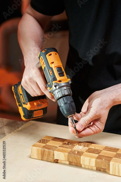 Fototapeta The hands of a Caucasian worker install a self-tapping screw into the bit of an electric screwdriver with a replaceable battery to screw it into a wooden part