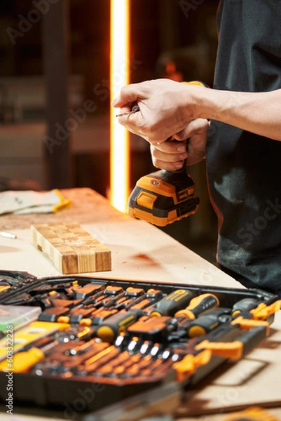 Fototapeta over a workbench with a bench tool, the hands of a worker tighten the drill into the chuck of an electric drill-driver with a replaceable battery