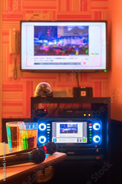 Fototapeta Two black microphones for singing songs on a corner of a table next to two drink glasses in a Japanese karaoke club room with a controller lit by backlit LEDs on buttons under a TV screen on the wall.