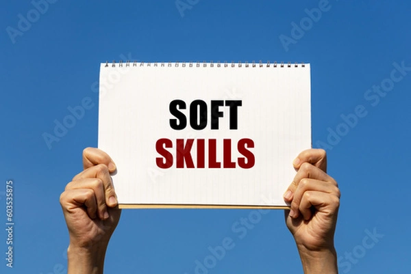Fototapeta Soft skills text on notebook paper held by 2 hands with isolated blue sky background. This message can be used as business concept about soft skills.