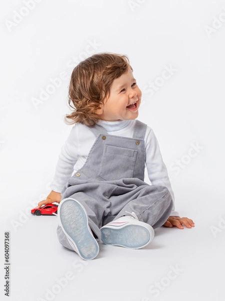 Fototapeta A cheerful laughing Caucasian toddler 2 years old with curly hair in a gray jumpsuit and a white turtleneck is playing with a car on the floor.