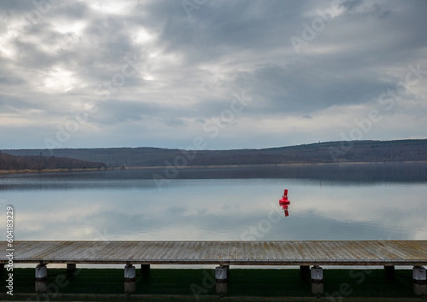 Fototapeta a scenic view of a lake with a wooden dock in the foreground. The sky is cloudy and there are trees in the background. In the water, there is also a red buoy visible.