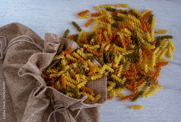 Obraz Uncooked multicolored Fusilli pasta spilled out of fabric bag over white wood background. top view.