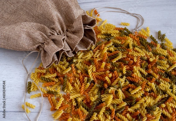 Obraz Uncooked multicolored Fusilli pasta spilled out of fabric bag over white wood background. top view.