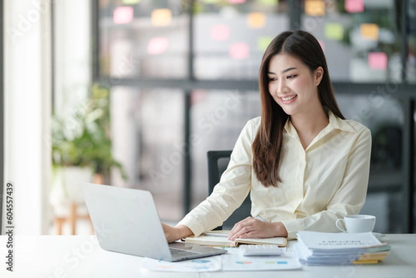 Fototapeta Photo of attractive woman sitting at desk working on computer looking at screen, smiling, wearing yellow shirt and glasses, typing on keyboard, holding a cup of coffee.