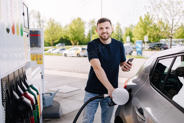 Fototapeta Bearded man refuelling car on gas station and looking into his smartphone. Man compares fuel prices 