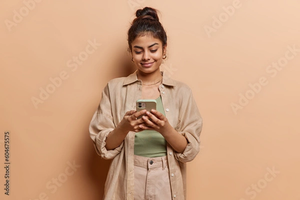 Fototapeta Indoor shot of beautiful charming young Indian woman uses smartphone for chatting online types message dressed in casual shirt and trousers browses social media isolated over brown background.