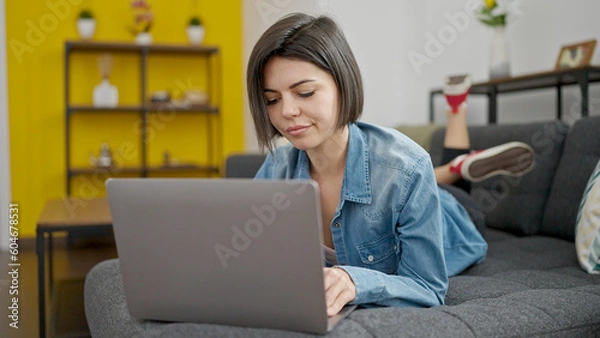 Obraz Young caucasian woman using laptop lying on sofa at home