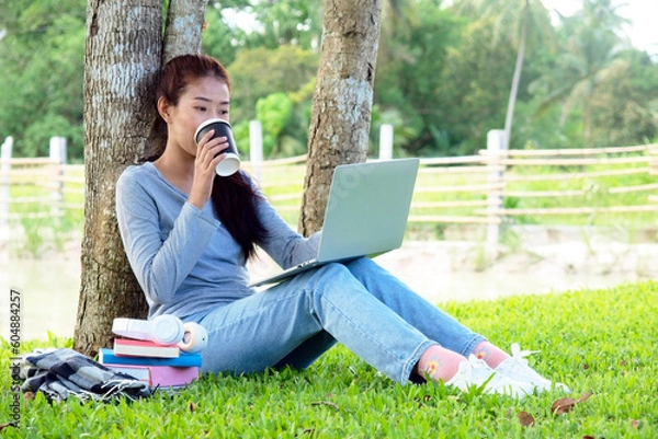 Fototapeta Asian student girl is studying  by laptop in nature with tree. Asian student is happy in study online and drinking coffee. Education in online life concept. Study Online in vacation.