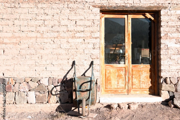 Fototapeta Frontal rural building with adobe brick wall entrance, double wooden door with crystal pane and light blue metal wheelbarrow stopped leaning on the warehouse wall