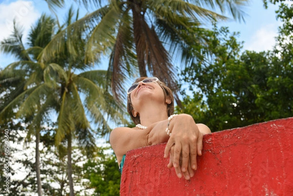 Obraz Portrait of senior woman 50-60 age looking up to the sky. Tourist standing near by the red wall on the beach. Concept - travel, retirement, active elderly people.