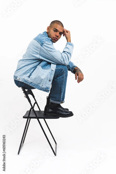 Fototapeta young and handsome Afro-American man in cool denim clothes sitting on the back of the chair and looking serious, studio shot vertical. High quality photo