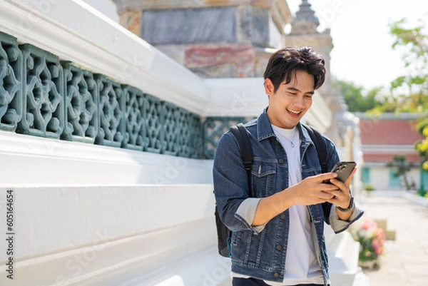 Obraz Portrait of Asian male traveler using a mobile phone on sidewalk of buddhist temple on street in Bangkok, Thailand, Southeast Asia - smart phone and internet for traveling concept