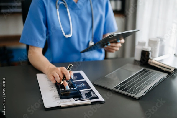Fototapeta Healthcare costs and fees concept.Hand of smart doctor used a calculator and smartphone, tablet for medical costs at hospital in morning light