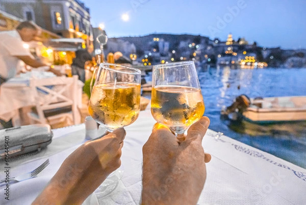 Fototapeta Raising hands holding a glass of white wine in front of a seaside village at a greek restaurant.