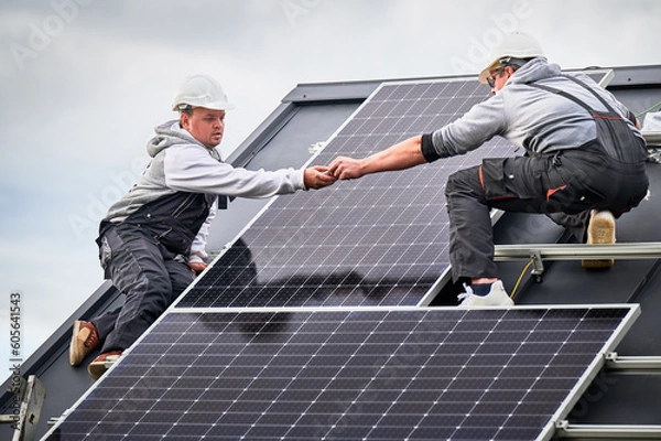 Fototapeta Workers building photovoltaic solar module station on roof of house. Men electricians in helmets installing solar panel system outdoors. Concept of alternative and renewable energy.