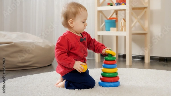 Fototapeta Little 1 year old baby boy sitting on floor and building colorful toy tower. Baby development, family playing games, making first steps, parenthood and care