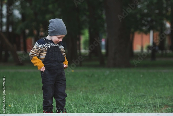 Fototapeta Portrait of a fashionable little boy outdoors. A happy child walks in the spring park. The kid is dressed in a fashionable sweater, sneakers and denim overalls.