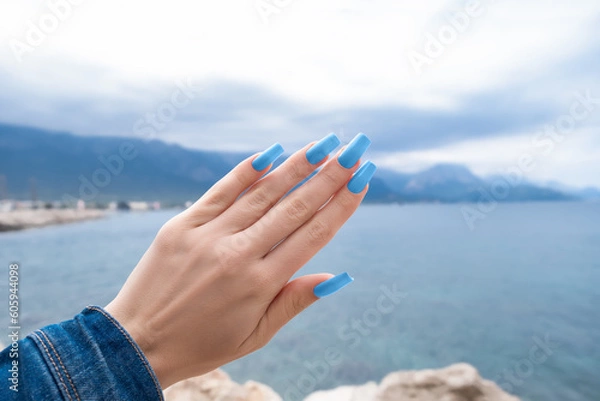 Fototapeta Female hand with blue long nail design. Glitter blue nail polish manicure. Hand with blue manicure on sea shore.
