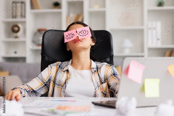 Obraz The concept of a lot of work. Tired Indian student girl sleeping with stickers on her eyes while sitting at the table. Distracted woman in home office.