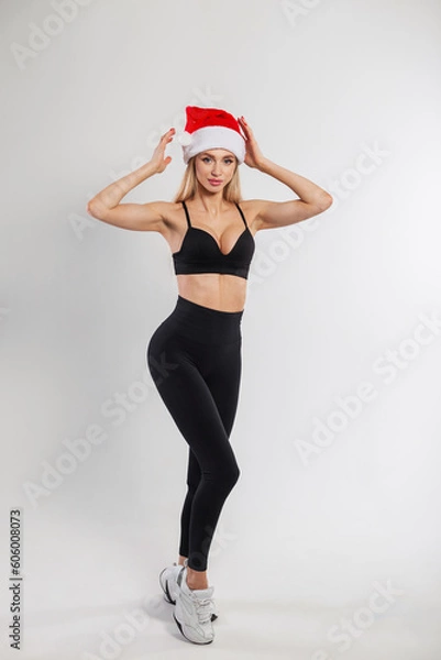 Obraz Beautiful young fitness lady with a slim body in black clothes with leggings and a bra with a red santa hat posing on a white background
