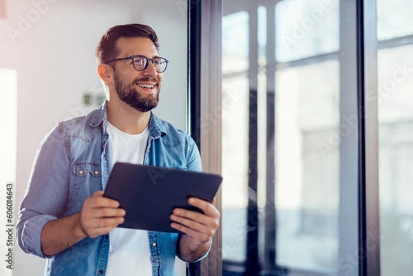 Obraz Smiling young businessman working online with digital tablet while standing by window.