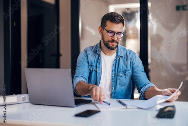 Obraz Satisfied young man with glasses sitting at a desk and doing paperwork at his workplace.