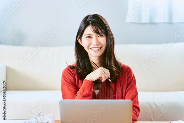 Obraz 自宅リビングのソファーの前でノートパソコンを前に笑顔でペンを持つカメラ目線の30代の1人の日本人女性