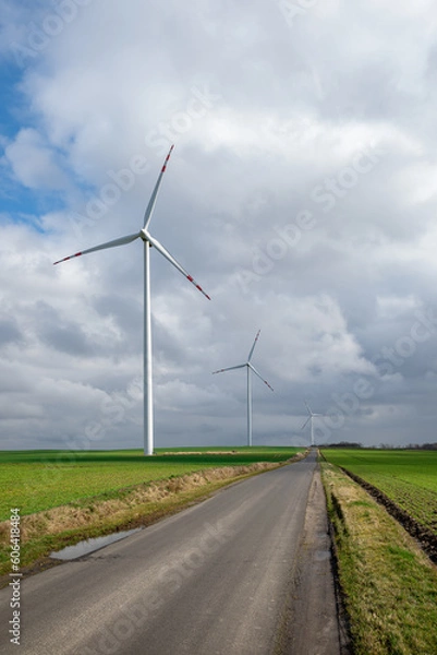Fototapeta Wind turbines against a cloudy sky standing in a green field with a tarmac road reaching to the horizon, Windmill for electricity generation