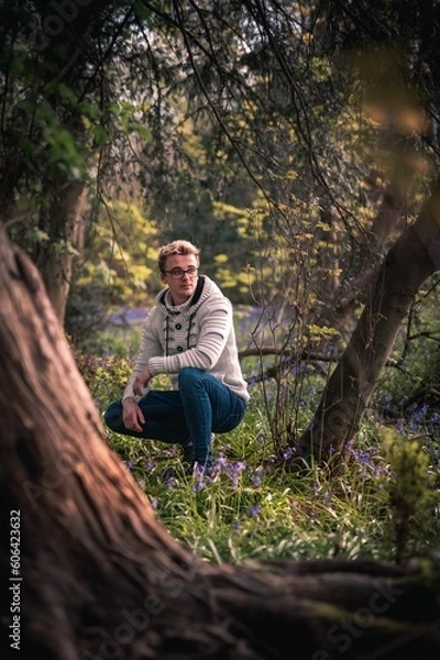 Fototapeta Vertical of an attractive Caucasian man posed squatting in a beautiful forest with wild flowers