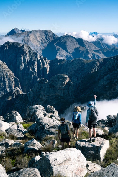 Fototapeta Hiking in the mountains with a group of hikers looking at the view of the mountains and clouds from the summit of a mountain. 