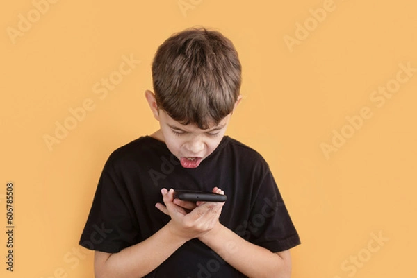 Fototapeta A young boy of 8 years looks at phone and shouts something, does not understand why the phone hangs during a difficult game, the concept of addiction to modern gadgets