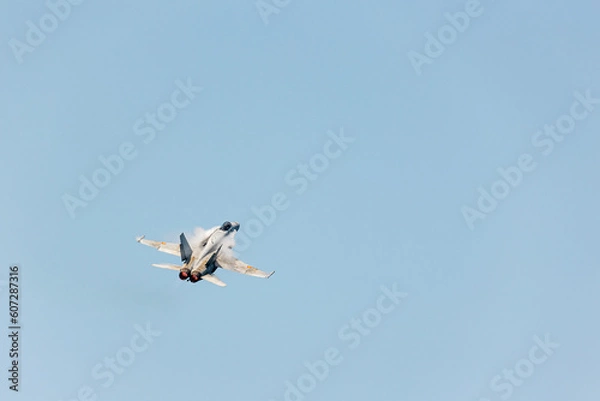 Fototapeta Top rear view of an F-18 jet fighter in Gijon Air show festival ascending at sonic speed in a clear blue sky with big condensation wakes in its wings and sunlight