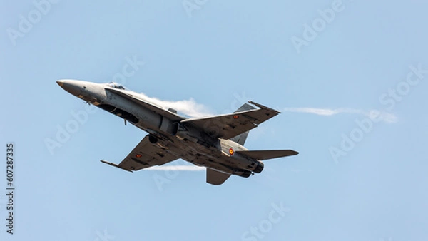 Fototapeta Front bottom view of an F-16 jet fighter in an Air show festival ascending at sonic speed in a clear blue sky with condensation big wakes in its wings