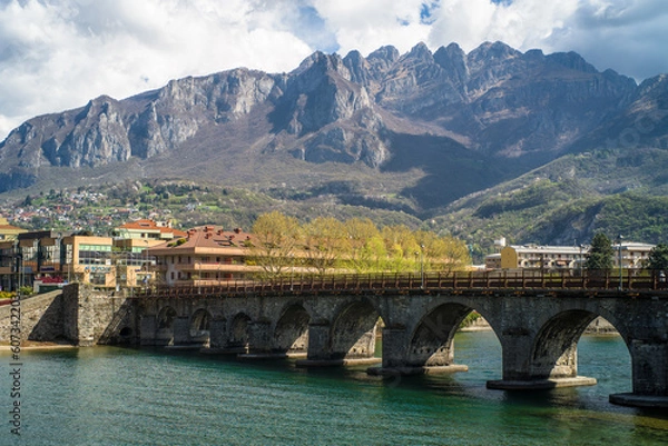Fototapeta Buildings of the city of Lecco on the banks of the river Adda. Bridges and an island with mountains in the background