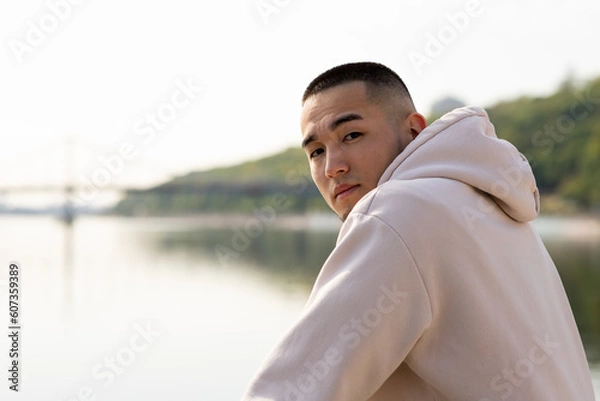 Fototapeta A young man stands by the river and looks at the camera over his shoulder. An Asian guy in a white hoodie poses for the camera against the backdrop of a blurred river.