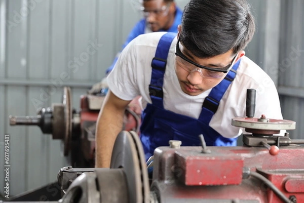 Fototapeta Multiethnic technicain mechanic or worker man in protective uniform using metal lathe machine operate polishing car disc brake at garage. Maintenance automotive and inspecting vehicle part concept