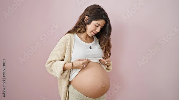 Fototapeta Young pregnant woman smiling confident showing belly over isolated pink background